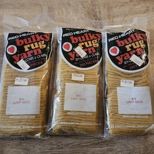 May include: Three packages of Red Heart bulky rug yarn in light gold, color 610. Each clear plastic bag contains 320 pieces of 100% Orlon acrylic fiber. The labels feature a red heart logo and the text Coats & Clark Inc., Stamford, Conn.