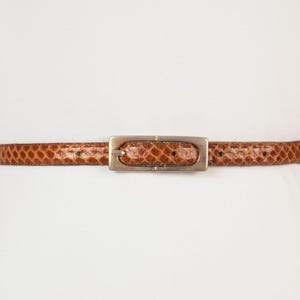Könnte beinhalten: Ein brauner Ledergürtel mit einer silbernen Schnalle. Der Gürtel ist aus einem strukturierten Material, das Schlangenleder ähnelt.