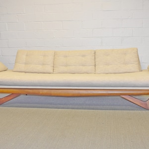 May include: A mid-century modern sofa with a light beige fabric upholstery and a wood frame. The sofa has a low profile and angled legs.