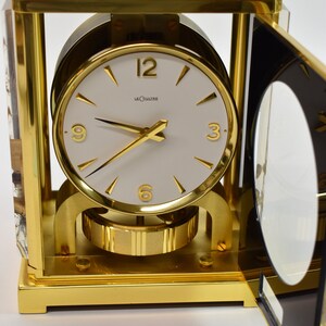 May include: A gold-toned, square-shaped clock with a white face and black hands. The clock has a glass top and a clear glass side panel. The clock face has the words "LeCoultre" printed on it.