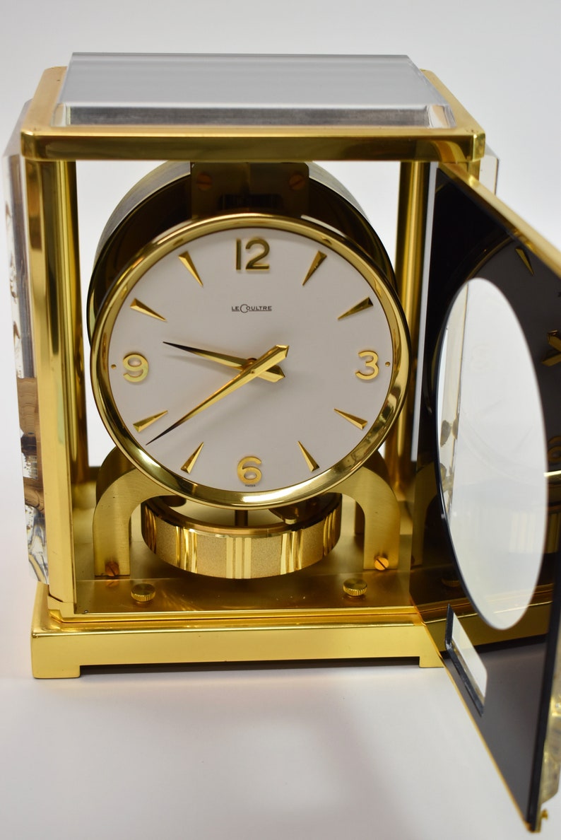 May include: A gold-toned, square-shaped clock with a white face and black hands. The clock has a glass top and a clear glass side panel. The clock face has the words "LeCoultre" printed on it.