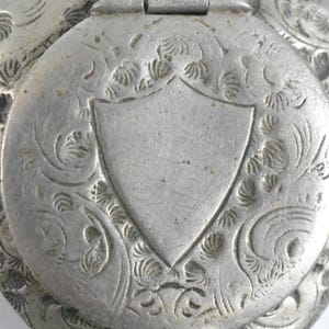 Puede incluir: Un relicario de metal plateado con una tapa circular con bisagras que presenta un escudo grabado en el centro. La superficie est&aacute; decorada con intrincados patrones florales y de volutas que rodean el escudo. El metal tiene un acabado mate y envejecido.