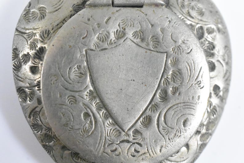 Puede incluir: Un relicario de metal plateado con una tapa circular con bisagras que presenta un escudo grabado en el centro. La superficie est&aacute; decorada con intrincados patrones florales y de volutas que rodean el escudo. El metal tiene un acabado mate y envejecido.