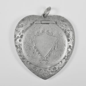 Puede incluir: Un relicario de metal plateado en forma de coraz&oacute;n con un intrincado patr&oacute;n floral en relieve que rodea un cartucho central liso en forma de escudo. El relicario incluye una secci&oacute;n superior con bisagras y una peque&ntilde;a argolla circular.