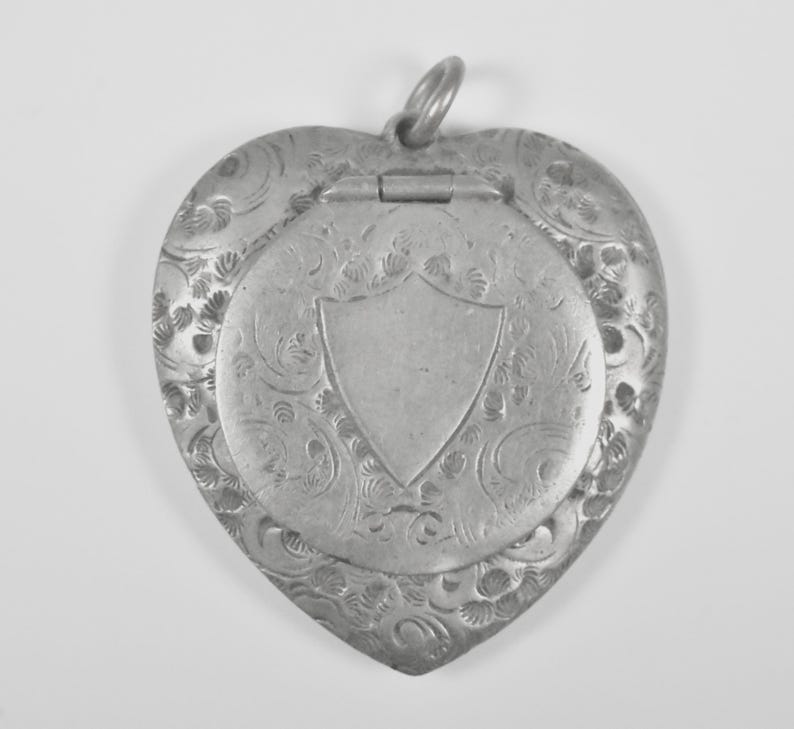 Puede incluir: Un relicario de metal plateado en forma de coraz&oacute;n con un intrincado patr&oacute;n floral en relieve que rodea un cartucho central liso en forma de escudo. El relicario incluye una secci&oacute;n superior con bisagras y una peque&ntilde;a argolla circular.