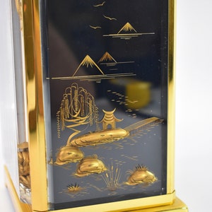 May include: A gold-framed box with a black background and a gold-colored Japanese landscape scene. The scene includes mountains, a lake, a bridge, and trees.