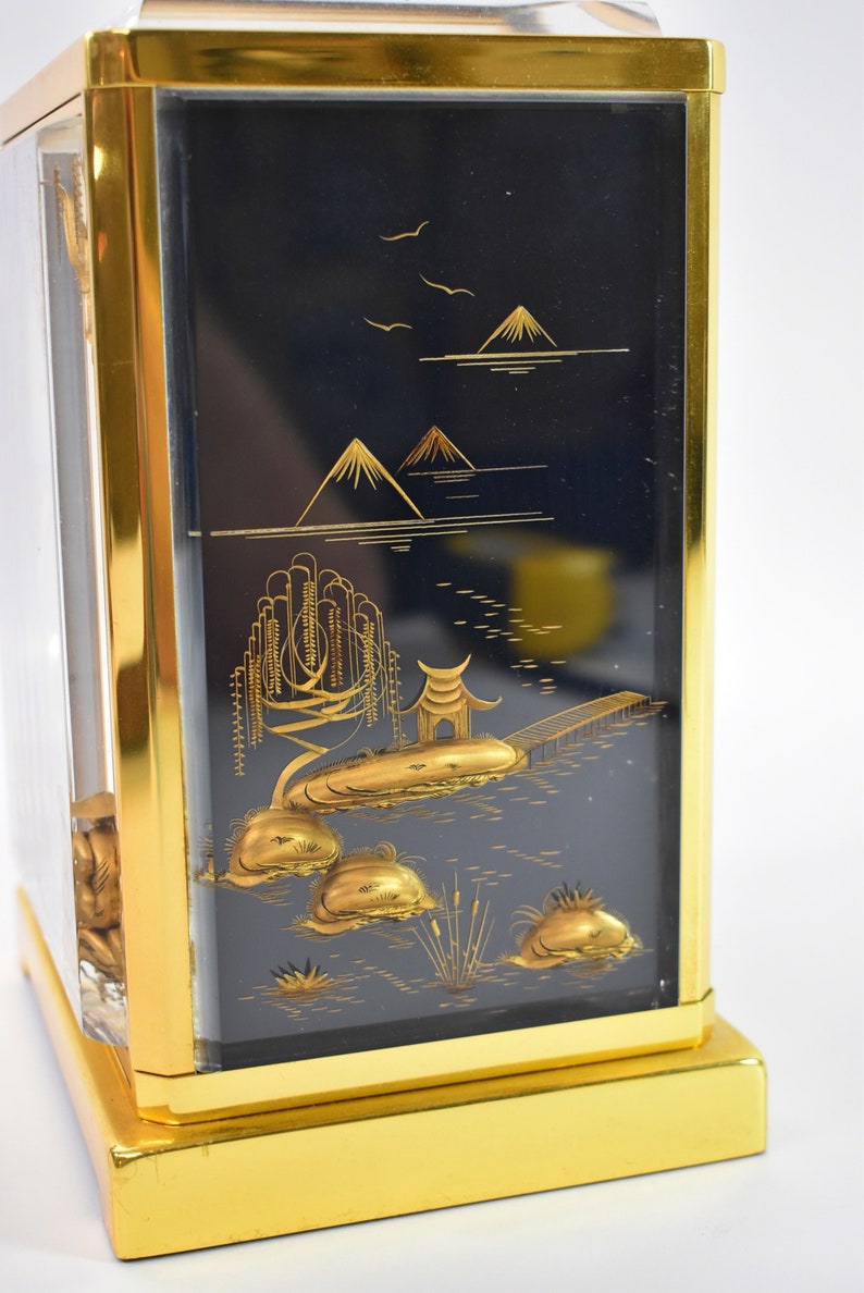 May include: A gold-framed box with a black background and a gold-colored Japanese landscape scene. The scene includes mountains, a lake, a bridge, and trees.