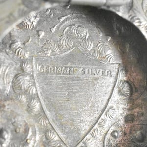 Puede incluir: Un objeto met&aacute;lico con acabado plateado que presenta un intrincado patr&oacute;n floral en relieve. Centrado en la superficie hay un emblema en forma de escudo con el texto GERMAN SILVER grabado horizontalmente a trav&eacute;s del centro del escudo.