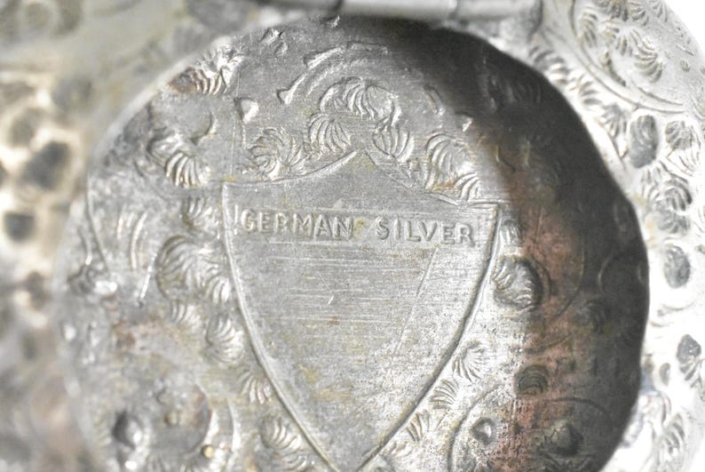 Puede incluir: Un objeto met&aacute;lico con acabado plateado que presenta un intrincado patr&oacute;n floral en relieve. Centrado en la superficie hay un emblema en forma de escudo con el texto GERMAN SILVER grabado horizontalmente a trav&eacute;s del centro del escudo.