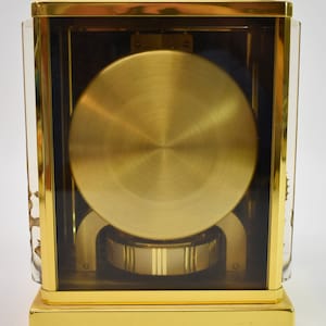 May include: A gold-toned clock with a clear glass case, revealing the intricate inner workings of the timepiece. The clock has a round, gold-colored face and a base with a polished finish.