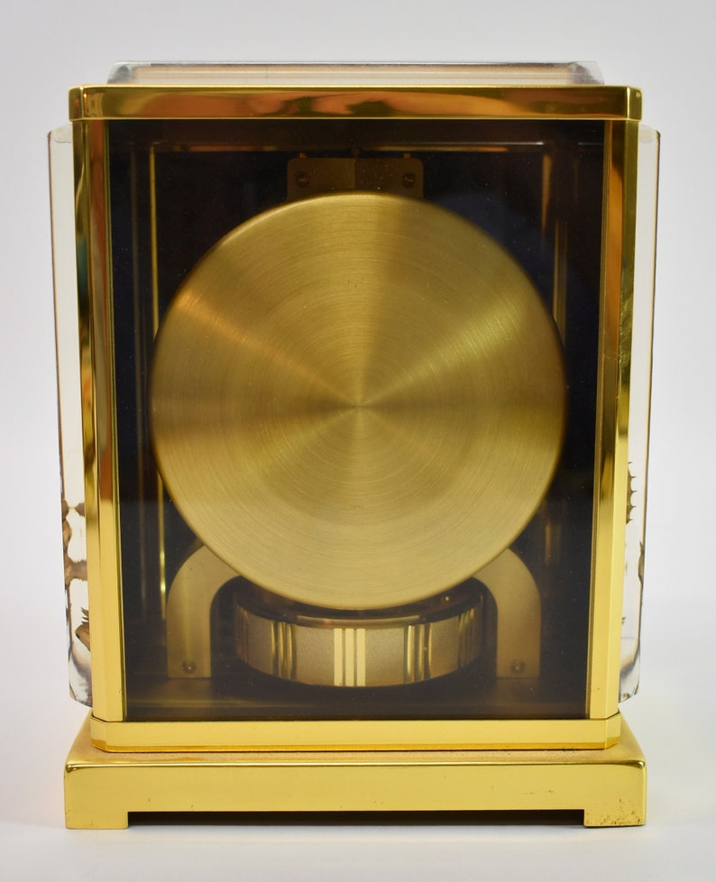 May include: A gold-toned clock with a clear glass case, revealing the intricate inner workings of the timepiece. The clock has a round, gold-colored face and a base with a polished finish.