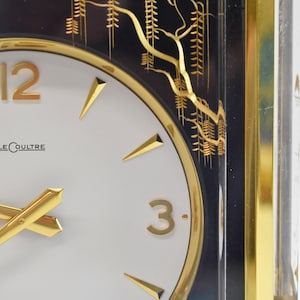 May include: A gold-framed clock with a white face and black Roman numeral hour markers. The clock has gold hands and a gold border around the face. The clock is set against a black background with a gold design of trees.