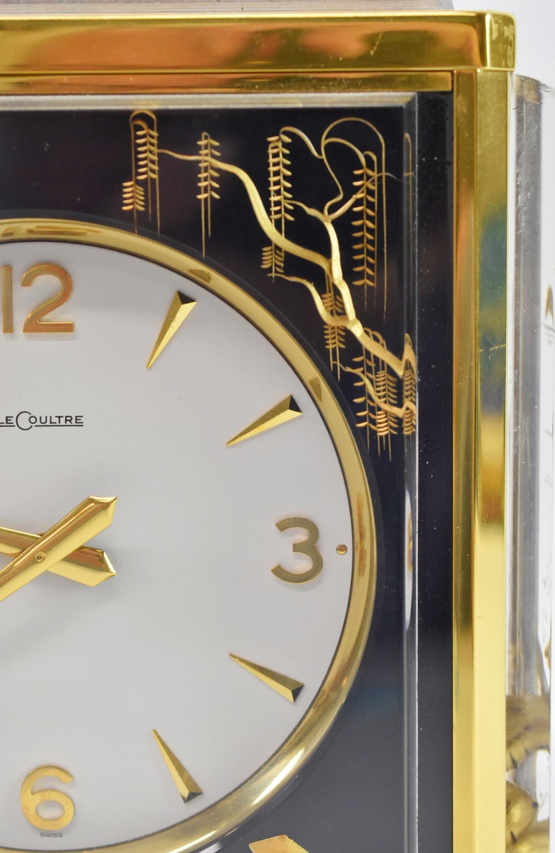 May include: A gold-framed clock with a white face and black Roman numeral hour markers. The clock has gold hands and a gold border around the face. The clock is set against a black background with a gold design of trees.