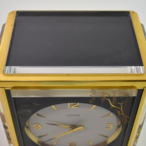 May include: A gold-toned clock with a black face and white hands. The clock has a glass front and a gold-toned frame. The clock face has the number 12 at the top, 9 on the left, and 3 on the right. The clock is made by the company "Movado".