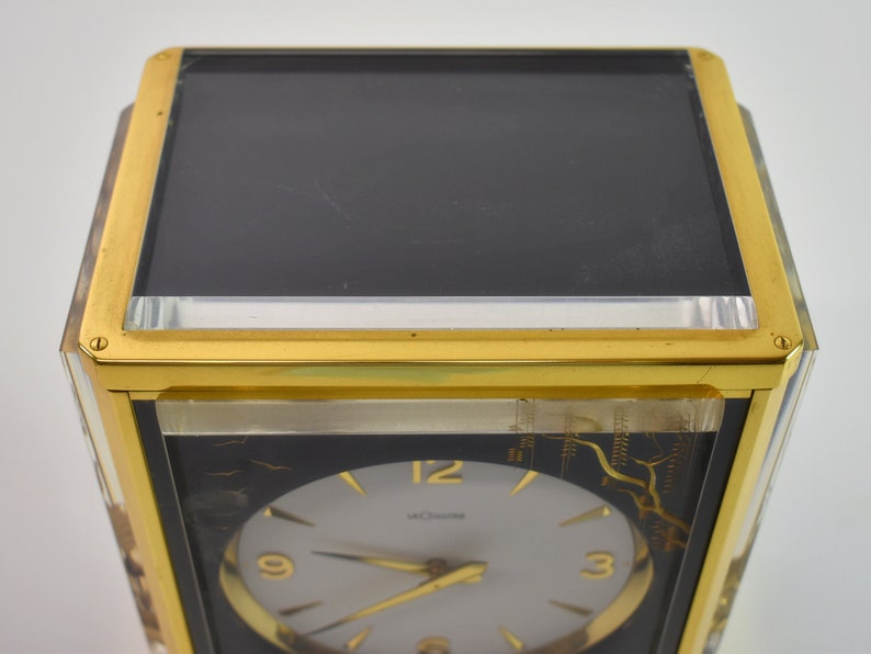 May include: A gold-toned clock with a black face and white hands. The clock has a glass front and a gold-toned frame. The clock face has the number 12 at the top, 9 on the left, and 3 on the right. The clock is made by the company "Movado".