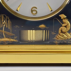 May include: A gold-toned mantle clock with a black face and gold accents. The clock face features Roman numerals and a gold hand. The clock is decorated with a scene of a fisherman in a boat on a lake. The clock is signed "Marina".