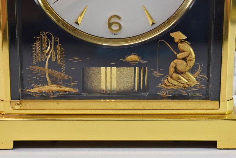 May include: A gold-toned mantle clock with a black face and gold accents. The clock face features Roman numerals and a gold hand. The clock is decorated with a scene of a fisherman in a boat on a lake. The clock is signed "Marina".