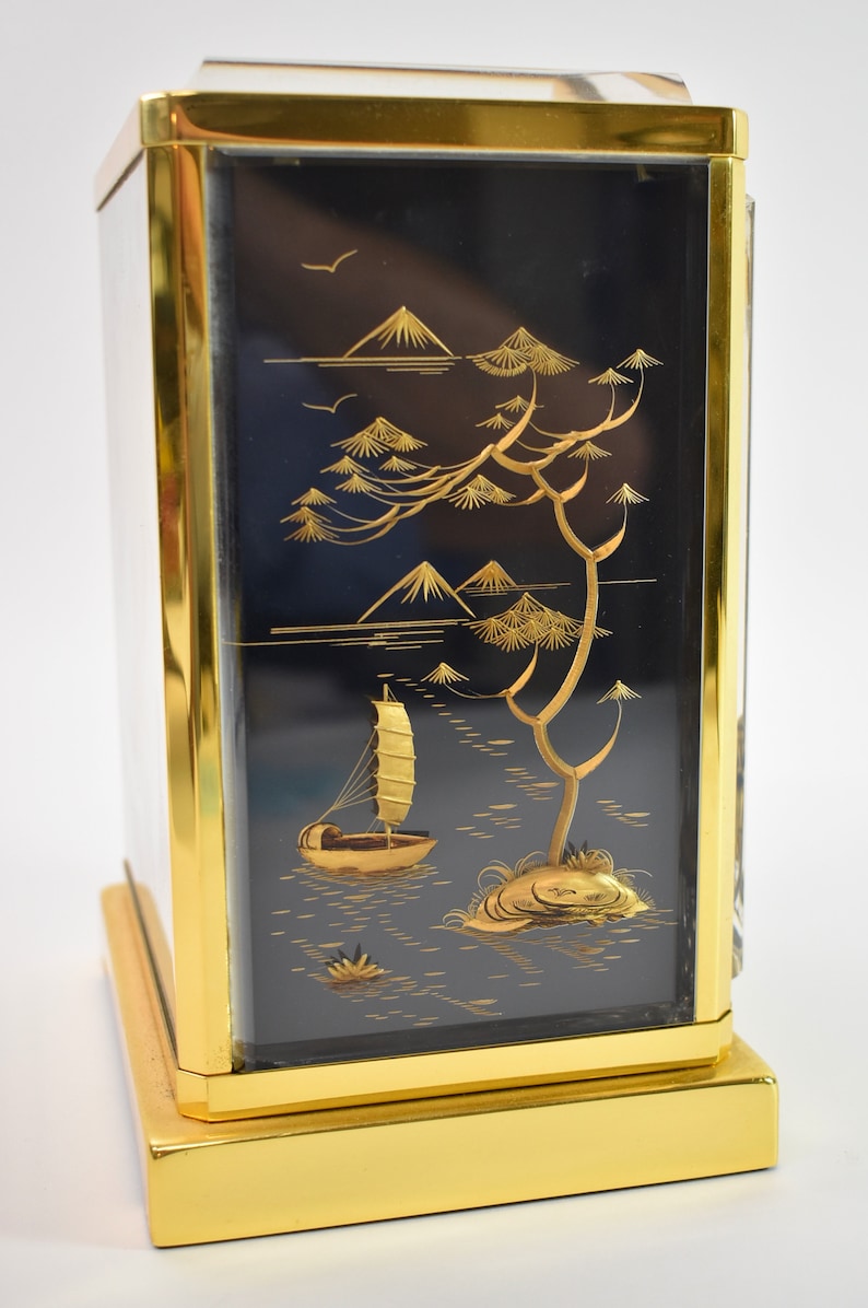 May include: A gold-framed box with a black background and a gold-colored scene of a sailboat on a lake with mountains and trees in the background.
