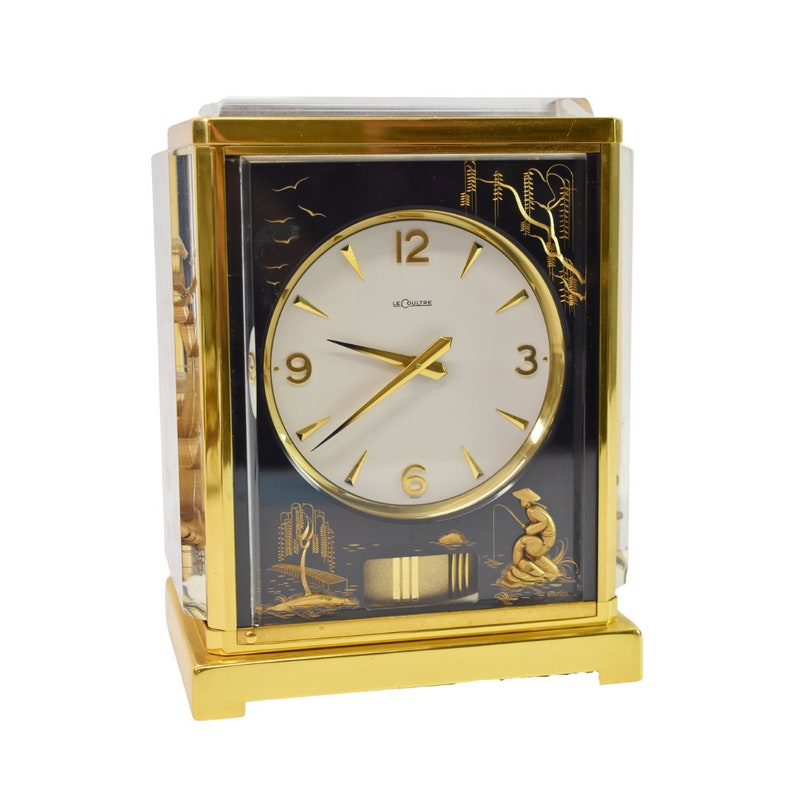 May include: A gold-toned LeCoultre clock with a white face and black Roman numeral markings. The clock has a black background with a gold-toned design of a fisherman and a boat.