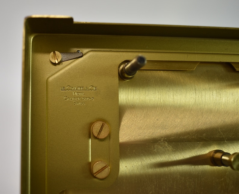 May include: Close-up of the back of a gold-colored watch movement. The movement is engraved with the text "Le Coultre & Cie Metal Caliber 15045 Swiss".