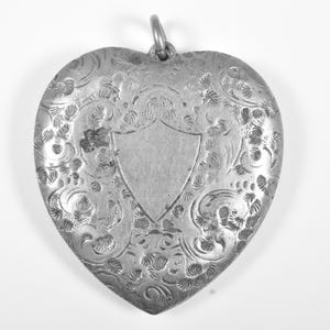 Puede incluir: Un colgante en forma de coraz&oacute;n de color plateado con un intrincado patr&oacute;n floral grabado en toda su superficie. El centro del colgante muestra un cartucho liso en forma de escudo. Una peque&ntilde;a anilla met&aacute;lica est&aacute; fijada en la parte superior.
