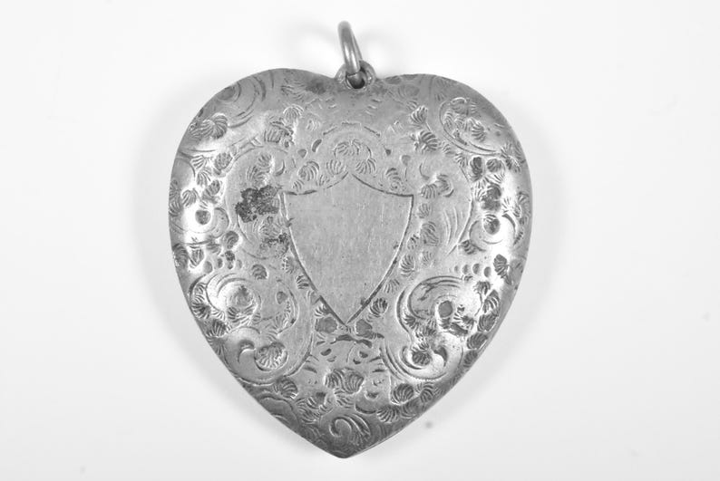 Puede incluir: Un colgante en forma de coraz&oacute;n de color plateado con un intrincado patr&oacute;n floral grabado en toda su superficie. El centro del colgante muestra un cartucho liso en forma de escudo. Una peque&ntilde;a anilla met&aacute;lica est&aacute; fijada en la parte superior.