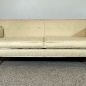 May include: A cream-colored sofa with tufted upholstery and dark wood legs. The sofa is sitting on a light gray carpet.