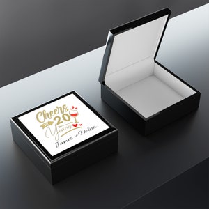 May include: Black wooden box with a white lid. The lid has a gold inscription that reads "Cheers to 20 Years James + Debra" with a red wine glass graphic.