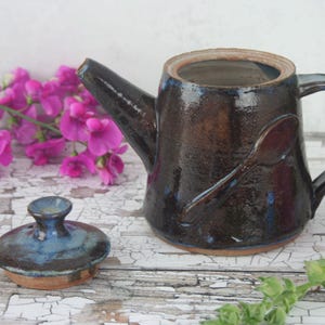 May include: A brown and blue ceramic teapot with a spoon design on the side. The teapot has a lid with a knob on top. The teapot is sitting on a white surface.