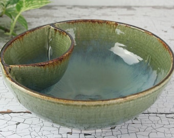 May include: A two-section ceramic serving bowl with a green and blue glaze. The bowl has a textured surface and a dark brown rim. The smaller section is attached to the larger bowl, creating a unique design.