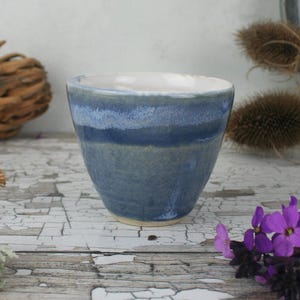 Könnte beinhalten: Ein Keramikbecher mit konischer Form und glatter, glänzender Glasur. Die Außenseite zeigt horizontale Bänder in tiefblauen, hellblauen und graublauen Tönen, während das Innere mit einer schlichten, einfarbig weißen Glasur versehen ist.