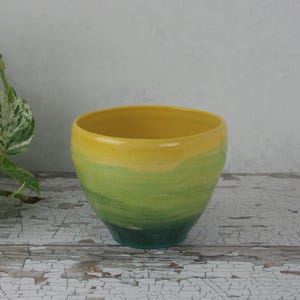 May include: A handmade ceramic bowl with a colour gradient. The top is yellow, fading into green and then a darker teal at the base. The bowl has a smooth, glossy finish and a rounded shape, suitable for decorative or functional use.