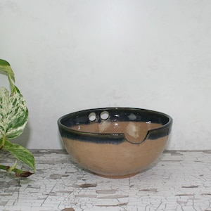 May include: A handmade ceramic bowl with a tan exterior and a dark blue rim. The bowl has three small holes near the rim and a small notch on the side. The bowl is sitting on a white, distressed wooden surface.