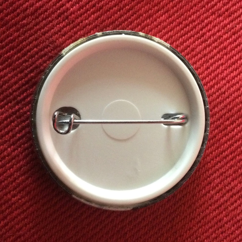 May include: The back of a round button with a silver pin and a white background.