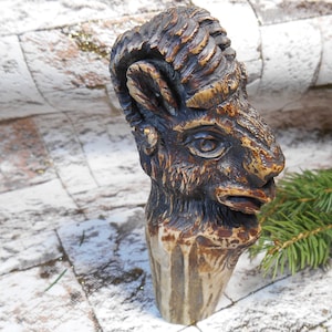 Puede incluir: Escultura detallada de una cabeza de carnero, que muestra texturas intrincadas y una paleta de colores naturales. La escultura presenta una base marrón oscuro con reflejos marrones claros y beige, capturando los rasgos del animal. La obra de arte se encuentra sobre un fondo texturizado.