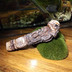 Puede incluir: Una pipa tallada en forma de calavera, marrón y beige, con detalles intrincados, apoyada sobre un trozo de musgo verde. La pipa tiene un diseño detallado y texturizado y está colocada sobre una superficie de madera. Es un artículo único hecho a mano.