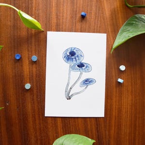 Op de afbeelding: Een aquarel van drie blauwe paddenstoelen met gedetailleerde doppen op een witte kaart. Het kunstwerk is omgeven door groene bladeren en kleine, kleurrijke, cirkelvormige objecten, tegen een donkere houten achtergrond.