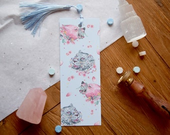 May include: A blue bookmark with a white tassel features a repeating pattern of pink and white animal faces with floral accents on a light blue background.