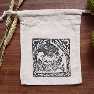 Może przedstawiać: Biała bawełniana torba ze sznurkiem, z czarno-białą ilustracją stworzenia z gwiazdami i koralikowym naszyjnikiem. Ilustracja jest oprawiona w czarną ramkę i wzory kwiatowe.