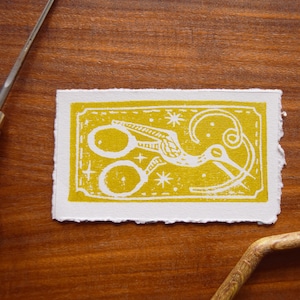 May include: A white linocut print on yellow paper featuring a stylized bird with a pair of scissors in its beak. The bird is surrounded by stars and a swirling design.