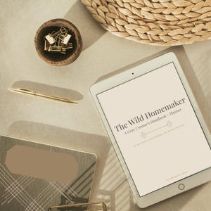 Könnte beinhalten: Eine Draufsicht mit einem weißen Tablet, das den Text "The Wild Homemaker" anzeigt, einem goldenen Stift, einer kleinen Holzschale mit goldenen Büroklammern und einem Weidenkorb. Ein Notizbuch und ein goldener Büroklammerhalter sind ebenfalls sichtbar.