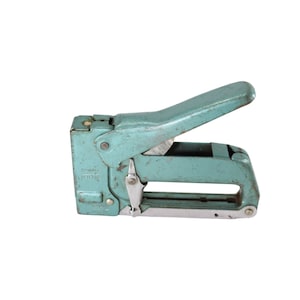 Puede incluir: Una grapadora vintage de metal azul con un mecanismo de carga de grapas de metal plateado. La grapadora tiene un mango y una palanca para operar la grapadora.