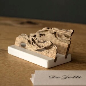 May include: A wooden business card holder with a natural wood finish and a white base. The holder is shaped like a wave and has a unique, organic design. The text "De Giolli" is printed on a stack of business cards.