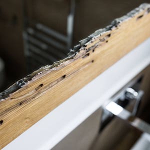 May include: Close-up of a wooden frame with signs of water damage. The frame is cracked and has dark brown stains. The frame is attached to a white surface.