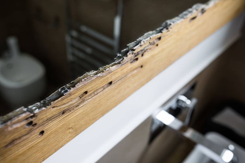 May include: Close-up of a wooden frame with signs of water damage. The frame is cracked and has dark brown stains. The frame is attached to a white surface.