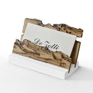 May include: A wooden business card holder with a natural wood finish and a white base. The holder has a unique design with intricate details and the text "De Zotti" on a card inside.