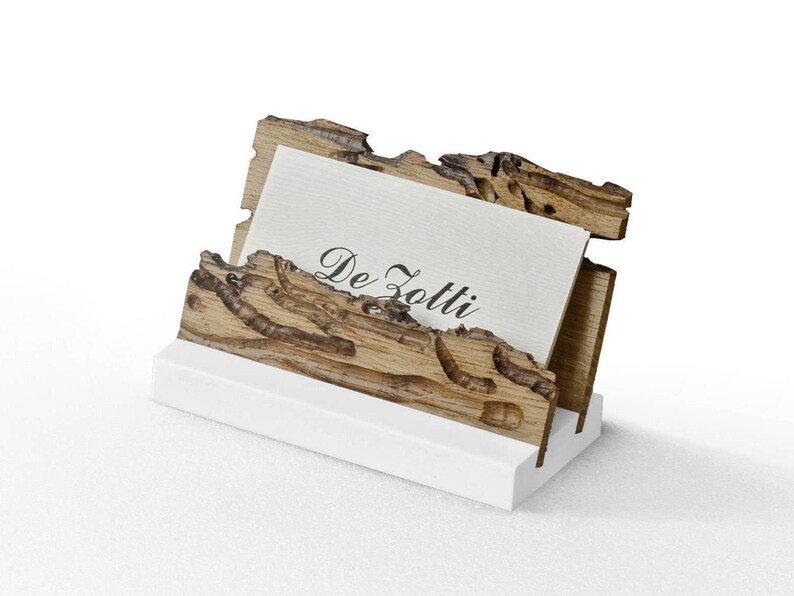 May include: A wooden business card holder with a natural wood finish and a white base. The holder has a unique design with intricate details and the text "De Zotti" on a card inside.