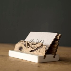 May include: A wooden business card holder with a natural wood finish. The holder is shaped like a piece of driftwood and has a white base. A business card is displayed in the holder with the text "De Folti" printed on it.
