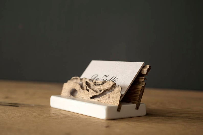 May include: A wooden business card holder with a natural wood finish. The holder is shaped like a piece of driftwood and has a white base. A business card is displayed in the holder with the text "De Folti" printed on it.