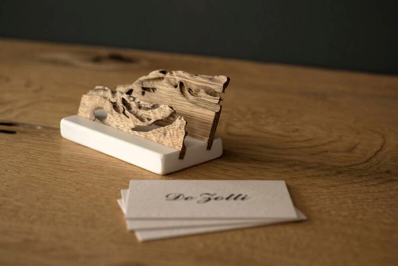 May include: A wooden business card holder with a natural wood finish and a white base. The holder is shaped like a wave and has a unique, organic design. The text "De Giolli" is printed on a stack of business cards.